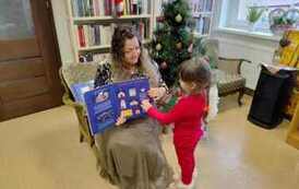 Ubrana na czerwono dziewczynka stoi przy bibliotekarce, kt&oacute;ra trzyma otwartą ksiażkę o świętach z okienkami i dziewczynka otwiera okienko w książce.