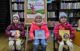 Trzy dziewczynki siedzą ma krzesełkach obok siebie trzymając nowe książki , za nimi stoi korkowa tablica z plakatem dotyczącym akcji Mała Książka Wielki Człowiek