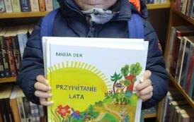 Chłopiec w jasnych włosach i w okularkach stoi pokazując swoją nową książkę.