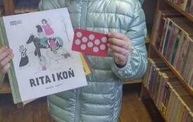 Uśmiechnięta dziewczynka w seledynowej kurtce i beżowej czapce stoi pokazując swoją nową książkę i kartę Młodego Czytelnika.