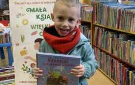 Chłopiec w blond włosach i w seledynowej kurtce stoi trzymając swoją nową książkę. Ma lekko rozchylone usta w uśmiechu. Za nim stoi tablica z plakatami dot. projektu.