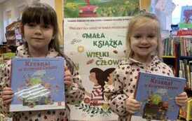 Dwie siostry stoją obok siebie i pokazują swoje nowe książki. Za nimi stoi korkowa tablica z plakatami dot. projektu. Jedna dziewczynka ma blond włosy, druga ciemne.