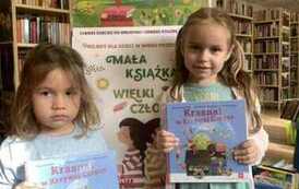 Dwie dziewczynki w jasnych włosach stoją obok siebie trzymając nowe książki. Za nimi stoi tablica z plakatami dot. projektu.