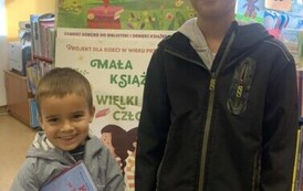 Mały uśmiechnięty chłopiec trzyma swoją nową książkę, obok stoi Jego starszy brat. Za nimi stoi tablica z plakatami dot. projektu Mała Książka Wielki Człowiek