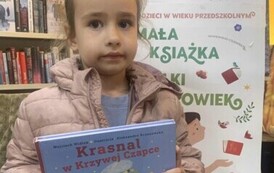 Dziewczynka w blond włosach i beżowej kurtce trzyma nową książkę, za Nią stoi tablica z plakatem dot. projektu Mała Książka Wielki Człowiek