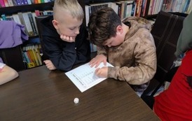 Dw&oacute;ch chłopc&oacute;w siedzi przy stole. Jeden z nich wpisuje zdanie na kartkę, a drugi patrzy. Obok kartki na stole leży kostka do gry.