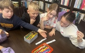 Dzieci siedzą przy stole blisko siebie i układają z rozsypanych wyraz&oacute;w zdania. Na stole leżą kredki i karteczki z rebusami.