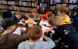 Dzieci siedzą przy stole i rozwiązują krzyż&oacute;wki wpisując hasła kredkami.