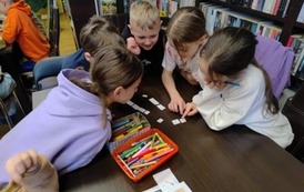 Dzieci siedzą przy stole blisko siebie i układają z rozsypanych wyraz&oacute;w zdania. Na stole leżą kredki i karteczki z rebusami.