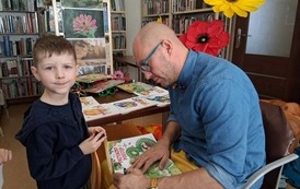 Chłopiec w granatowej bluzie stoi przy Panu Poecie, kt&oacute;ry na kolanach podpisuje mu zakupioną książeczkę.