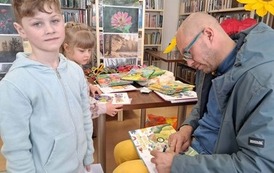 Chłopiec w blond włosach stoi przy Panu Poecie, kt&oacute;ry podpisuje Mu zakupioną książkę. Za nim stoi dziewczynka.