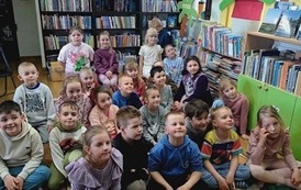 Grupa dzieci siedzi na dywanie i patrzy w stronę bibliotekarki prowadzącej zajęcia