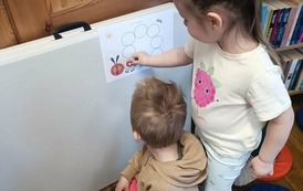 Dziewczynka stoi przy kartce na kt&oacute;rej jest gąsienica i przylepia w odpowiednie miejsce jabłko. Obok niej siedzi chłopiec.