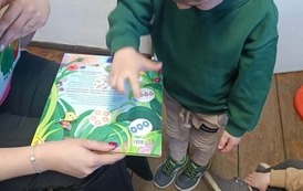 Chłopiec w jasnych włosach i zielonej bluzie stoi przy bibliotekarce, kt&oacute;ra trzyma książkę o owadach i paluszkiem pokazuje jedzenie dla gąsienicy