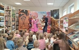 Dzieci siedzą na dywanie i patrzą na Zosię w r&oacute;żowej sukience i obok stojącego Feliksa trzymającego dużego pluszowego pieska.