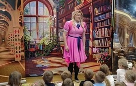Kurtyna przedstawiająca bibliotekę, stoi filmowa Zosia w r&oacute;żowej sukience i w okularach i patrzy w stronę publiczności dziecięcej.