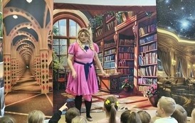 Kurtyna przedstawiająca bibliotekę, stoi filmowa Zosia w r&oacute;żowej sukience i w okularach i patrzy w stronę publiczności dziecięcej. Ma lekko rozłożone ręce.