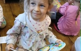 Dziewczynka w blond włosach siedzi na podłodze i ogląda książeczkę. Za nią widać inne dzieci.