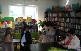 Pani Ania siedzi przy stole w bibliotece, obok siedzi w przebraniu duża maskotka jeża, a obok stoją dzieci w przebraniu czarodzieja  i wr&oacute;żki