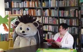 Pani Ania siedzi przy stole w bibliotece i czyta bajkę, obok siedzi duża maskotka w przebraniu. W tle regały z ksiązkami.