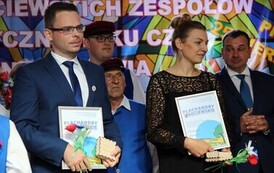 Stoją: Kobieta i kliku mężczyzn, trzymają obrazki z tytułem Plachandry Kociewskie, pudełeczka i sztuczne kwiaty.