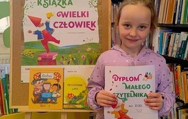Uśmiechnięta dziewczynka w blond włosach stoi w bibliotece w Pinczynie przy tablicy z plakatem projektu i pokazuje sw&oacute;j nowy dyplom Małego Czytelnika.