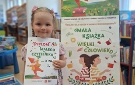 Uśmiechnięta dziewczynka w blond warkoczykach stoi w bibliotece w Zblewie przy tablicy z plakatami Mała Książka Wielki Człowiek i pokazuje sw&oacute;j dyplom Małego Czytelnika