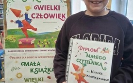 Uśmiechnięty chłopiec w okularach stoi w bibliotece w Zblewie przy tablicy z plakatami Mała Książka Wielki Człowiek i pokazuje sw&oacute;j dyplom