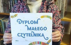 Uśmiechnięta dziewczynka w blond włosach stoi w bibliotece w Borzechowie i pokazuje sw&oacute;j nowy dyplom Małego Czytelnika.