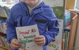 Chłopiec w blond włosach i niebieskiej kurtce stoi w bibliotece w Zblewie przy tablicy z plakatami Mała Książka Wielki Człowiek i trzyma sw&oacute;j nowy dyplom.