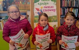 Trzy dziewczynki siostry stoją w bibliotece w Pinczynie przy korkowej tablicy z plakatem Mała Książka Wielki Człowiek i pokazują swoje dyplomy.