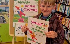 Uśmiechnięty chłopiec stoi w bibliotece w Pinczynie przy korkowej tablicy z plakatem Mała Książka Wielki Człowiek i trzyma sw&oacute;j nowy dyplom.