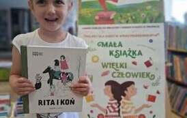 Uśmiechnięta dziewczynka w blond wlosach pokazuje swoja nową książkę pt Rita i koń. Za nią stoi tablica z plakatami projektu Mała Książka Wielki Człowiek