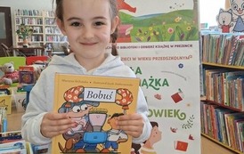 Uśmiechnięta dziewczynka w ciemnych warkoczach pkazuje swoją nową książkę pt Bobuś. Za nią stoi tablica z plakatami projektu Mała Książka Wielki Człowiek