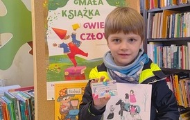 Chlopiec w blond włosach pokazuje swoją nową książkę i kartę Małego Czytelnika. Za nim stoi korkowa tablica z plakatem projektu Mała Książka Wielki Człowiek