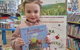 Dziewczynka w blond włosach trzyma swoją nową książkę, za nią stoi tablica z plakatami projektu Mała Książka Wielki Człowiek