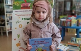 Dziewczynka w blond włosach trzyma swoją nową książkę, za nią stoi tablica z plakatami projektu Mała Książka Wielki Człowiek