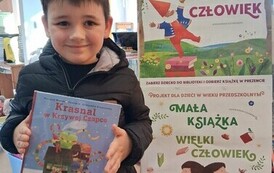 Chłopiec w ciemnych włosach i w czarnej kurtce pokazuje swoją nową książkę. Obok stoi tablica z plakatami projektu Mała Książka Wielki Człowiek