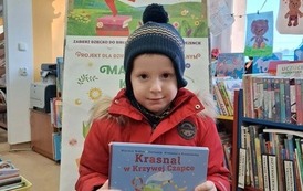 Chłopiec w czerwonej kurtce trzyma swoją nową książkę, za nim stoi tablica z plakatami projektu Mała Książka Wielki Człowiek