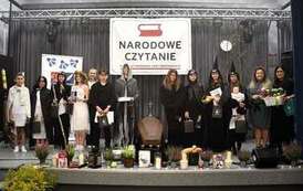 Grupa uczestnik&oacute;w Narodowego Czytania Dziad&oacute;w Adama Mickiewicza  stoi na scenie w strojach nawiązujących do dramatu. W tle napis "Narodowe Czytanie", a scenę ozdabiają dekoracje i rekwizyty nawiązujące do obrzędu dziad&oacute;w. Wydarzenie odbywa się w sali widowiskowej GOK