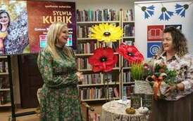 Kierowniczka z koszykiem kwiat&oacute;w w ręku stoi przy pisarce dziękując za spotkanie. W tle baner z logiem biblioteki, dekoracje kwiatowe i duży ekran z wyświetlanym plakatem ze spotkania.