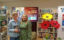 Czytelniczka z zakupioną książką stoi przy pisarce pozując. W tle duży ekran z wyświetlanym plakatem ze spotkania i dekoracje kwiatowe.