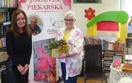 Bibliotekarka stoi obok pisarki trzymającej stroik jesienny. W tle baner promujący książkę Jolaanty Piekarskiej.