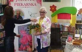 Bibliotekarka wręcza pisarce stroik jesienny w podziękowaniu za spotkanie. W tle baner promujący książkę pt " Warto. Naprawdę warto"