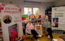 Pisarka siedzi z przodu wraz z mężem przy stoliku. Po bokach stoją banery promujące Centrum Szkolenia Panorama i książkę Jolanty Piekarskiej.