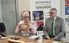 Pisarka siedzi obok męża przy stole z poczęstunkiem, trzyma w ręku koszyk, w kt&oacute;rym są na karteczkach ciekawe sentencje, myśli do losowania dla czytelnik&oacute;w. Za nimi baner promujący Centrum Szkolenia Panorama