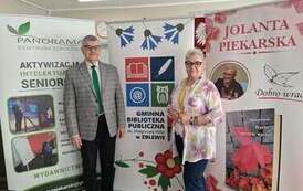 Pisarka i Jej mąż stoją obok siebie na tle baner&oacute;w promujących Centrum Szkolenia Panorama, Gminną Bibliotekę Publiczną w Zblewie oraz książki Jolanty Piekarskiej.