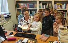 Dzieci stoją przy biurku, bibliotekarka pomaga dziewczynce przyłożyć książkę do czytnika.