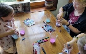 Trzy dziewczynki przy stole wsypują do plastikowych kubeczk&oacute;w kolorowe proszki, na stole leżą też kolorowe pisaki i nożyczki.