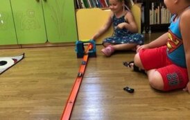 Chlopiec i dziewczynka siedzą na podłodze bawiąc się autami. Na podlodze leży rozłożony tor samochodowy.